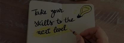 A photo of a hand holding a pen with a drawing of a light bulb, ‘Take Your Skills to the Next Level’ written across a page