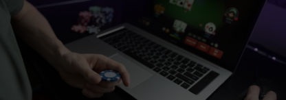 A photo of a man holding a poker chip in front of a laptop with a blurred screen showing an online poker table with players 