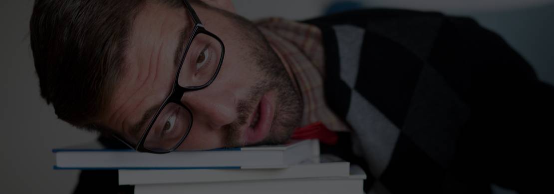 geeky looking guy collapsing on a pile of books he's studying in front of his computer